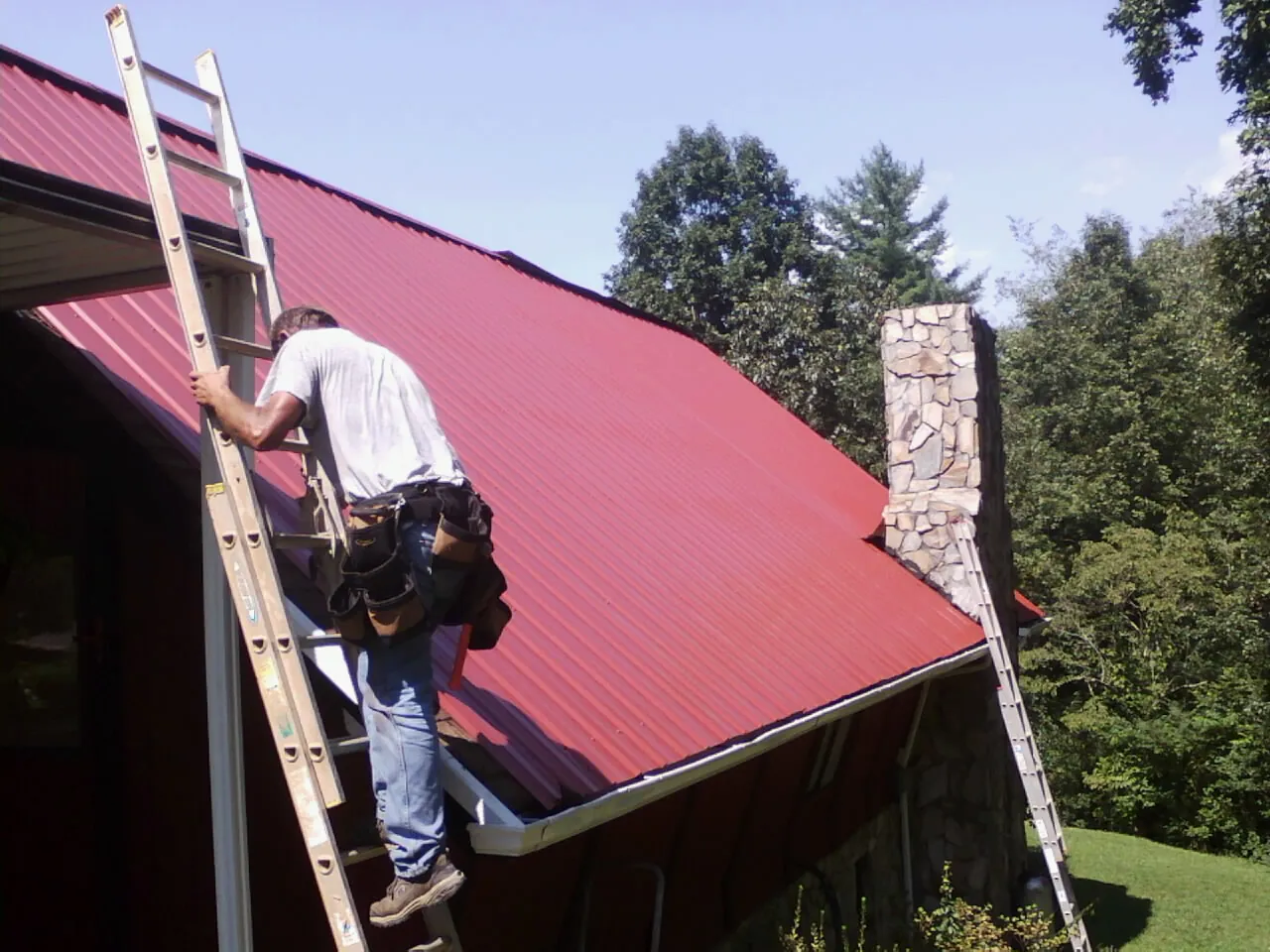 Roofer on a ladder beside a red metal roof and stone chimney — Soffit & Fascia Repair by Roofing Brothers in North Chatham